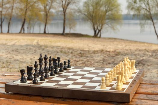 Chess board with chess pieces on wooden bench with river embankment background. Chess game with wooden chess pieces