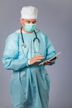 Medical doctor dressed in a medical gown with stethoscope and face mask holding a tablet. Gray background