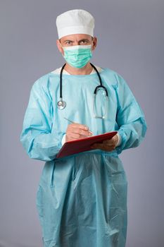 Medical doctor dressed in a medical gown with stethoscope and face mask holding a pen and writing in a notebook. Gray background