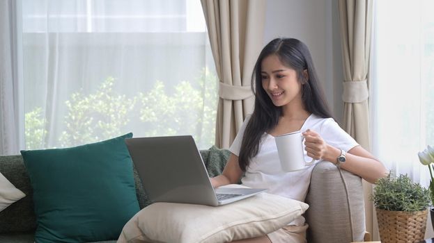 Casual young asian woman resting on couch and using laptop, surfing internet, checking email.