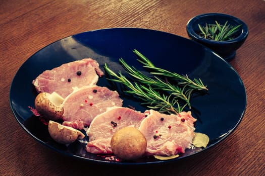 Pieces of raw pork steak with spices and herbs rosemary, mushrooms, salt and pepper on a black plate