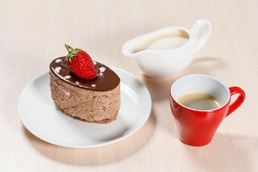 Chocolate cake with strawberry on the white plate, a cup of coffee and a gravy boat with milk on a wooden table