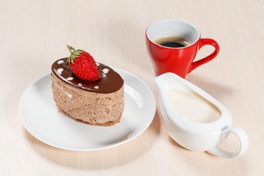 Chocolate cake with strawberry on the white plate, a cup of coffee and a gravy boat with cream on a wooden table