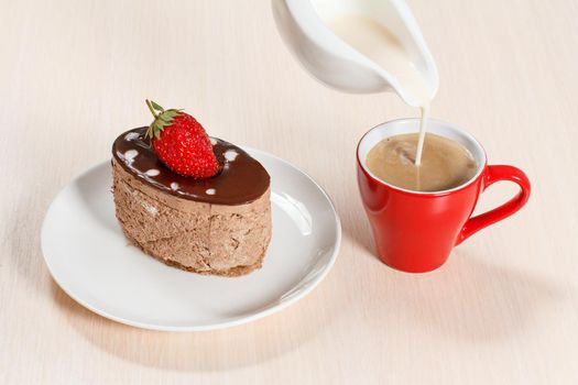 Chocolate cake with strawberry on the white plate, a cup of coffee in which milk is added from a gravy boat