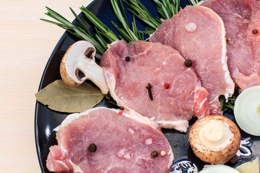 Pieces of raw pork steak with spices and herbs rosemary, mushrooms, onion, bay leaf, salt and pepper on a black plate and white wooden desk, top view