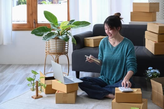 Young asian woman using calculator and preparing products for deliver to customers. SME marketing concept.