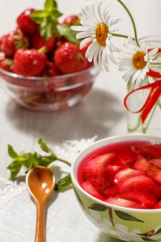 Cherry jelly with strawberry pieces in the bowl with strawberries in a bowl and chamomiles on the background