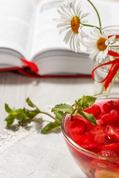 Cherry jelly with strawberry pieces in the glass bowl topped mint leaves with opened book and chamomiles on the background