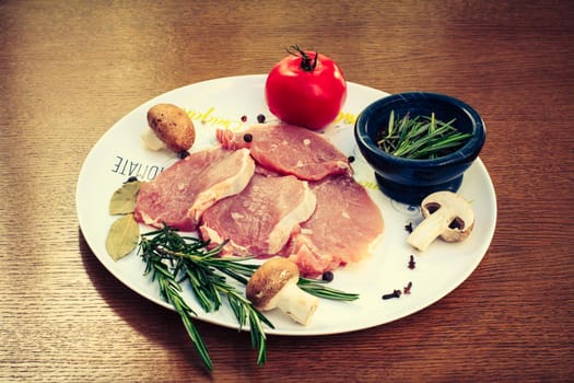 Pieces of raw pork steak with spices and herbs rosemary, mushrooms, tomato, salt and pepper on a white plate