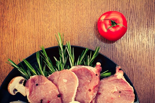 Pieces of raw pork steak with spices and herbs rosemary, mushrooms, tomato, salt and pepper on a black plate, top view