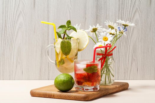 Cold refreshing summer lemonade mojito with lemon slices and mint in a tall glass and strawberry lemonade with mint in a short one on wooden cutting board