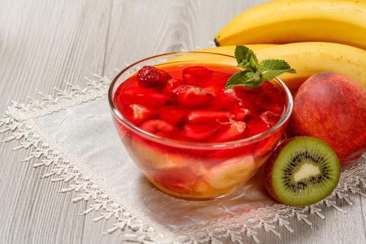 Cherry jelly with strawberry pieces in the glass bowl topped mint leaves, nectarine, kiwi and banana on the white napkin