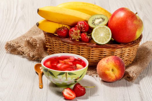 Cherry jelly with strawberry pieces in the bowl, fresh strawberries, nectarine, lime, kiwi, banana, apple in a wicker basket on a wooden table