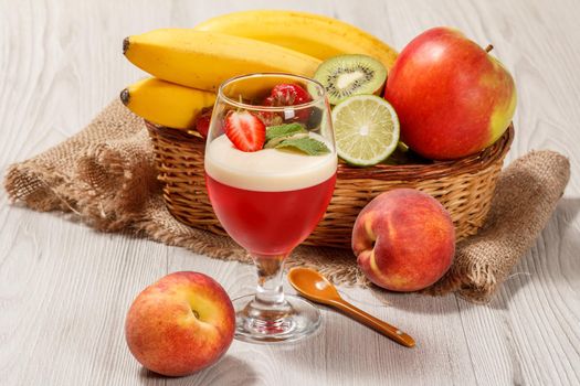 Cherry and milk jelly with strawberry piece in the glass topped mint leaves, fresh strawberries, nectarine, lime, kiwi, banana, apple in a wicker basket on a wooden table