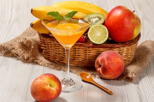 Orange jelly in the glass topped mint leaves, fresh strawberries, nectarine, lime, kiwi, banana, apple in a wicker basket on a wooden table