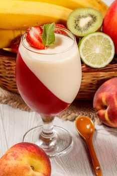 Cherry and milk jelly with strawberry piece in the glass topped mint leaves, fresh strawberries, nectarine, lime, kiwi, banana, apple in a wicker basket on a wooden table