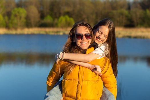 Playful mother giving daughter piggy back ride at spring lake shore. Both laughing and look happy. Spring in lake background. Closeup.