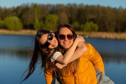 Playful mother giving daughter piggy back ride at spring lake shore. Both laughing and look happy. Spring in lake background. Closeup.