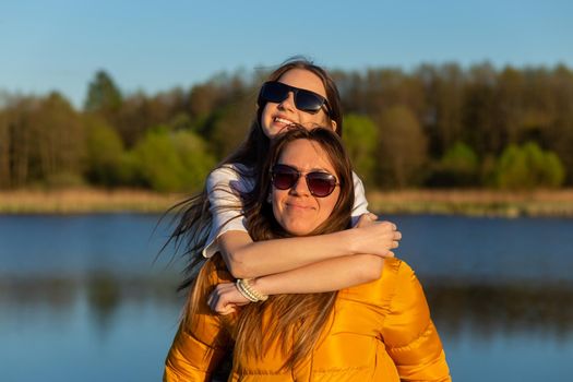 Playful mother giving daughter piggy back ride at spring lake shore. Both laughing and look happy. Spring in lake background. Closeup.