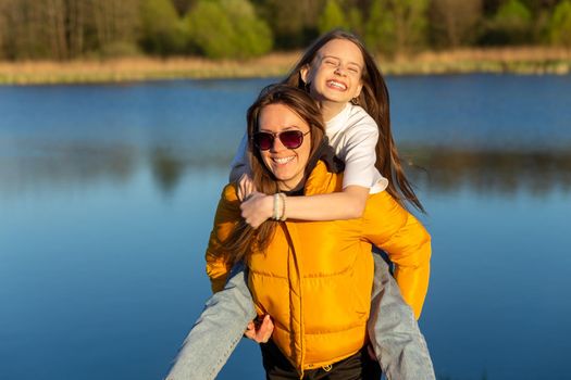 Playful mother giving daughter piggy back ride at spring lake shore. Both laughing and look happy. Spring in lake background. Closeup.