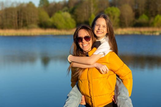 Playful mother giving daughter piggy back ride at spring lake shore. Both laughing and look happy. Spring in lake background. Closeup.