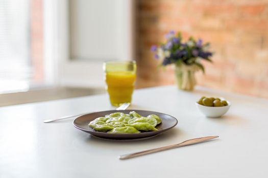 Traditional russian and ukrainian vegetarian green vareniki dumplings on a white plate, with olives and orange juice. Dough colored with spinach juice. Brick wall and window as the background.
