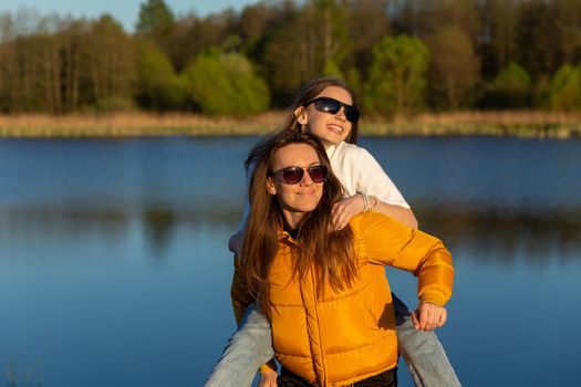 Playful mother giving daughter piggy back ride at spring lake shore. Both laughing and look happy. Spring in lake background. Closeup.