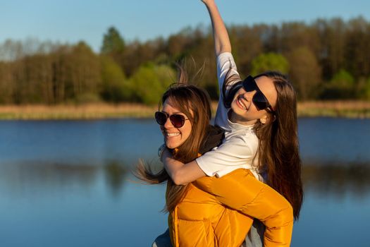 Playful mother giving daughter piggy back ride at spring lake shore. Both laughing and look happy. Spring in lake background. Closeup.