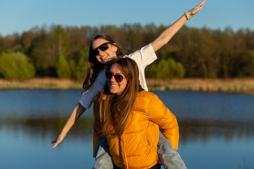 Playful mother giving daughter piggy back ride at spring lake shore. Both laughing and look happy. Spring in lake background. Closeup.