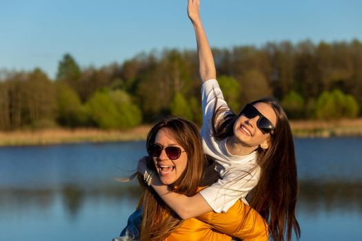 Playful mother giving daughter piggy back ride at spring lake shore. Both laughing and look happy. Spring in lake background. Closeup.