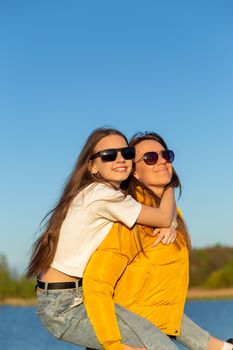 Playful mother giving daughter piggy back ride at spring lake shore. Both laughing and look happy. Spring in lake background. Closeup.