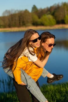 Playful mother giving daughter piggy back ride at spring lake shore. Both laughing and look happy. Spring in lake background. Closeup.