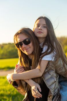 Playful mother giving daughter piggy back ride at green field. Both laughing and look happy. Spring in forest background. Closeup.