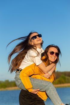 Playful mother giving daughter piggy back ride at spring lake shore. Both laughing and look happy. Spring in lake background. Closeup.