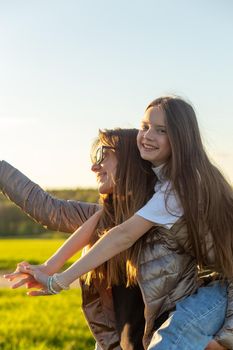 Playful mother giving daughter piggy back ride at green field. Both laughing and look happy. Spring in forest background. Closeup.