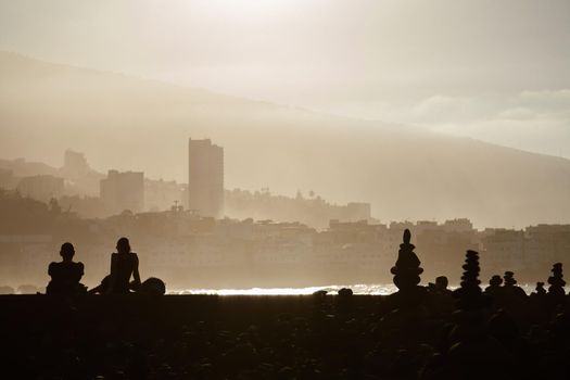People are sitting on the beach on a sunny evening and looking at the sea. In the background the coast and buildings in a haze of water. Tenerife. Puerto de la Cruz. Playa Jardin.