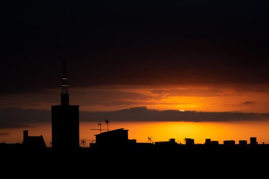 Sun is hiding behind the clouds at sunset, roof top view of an old church tower.. Summer sunrise or sunset. Dark orange sky color tones. Old town of Badalona, Spain