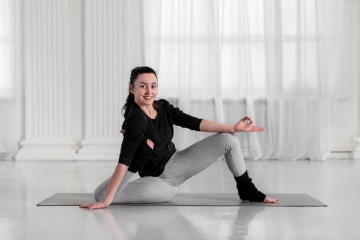 Beautiful asian young woman practicing yoga. Happy and smiling during workout, indoors, doing doing variation of yoga asana on gray mat in white hall.