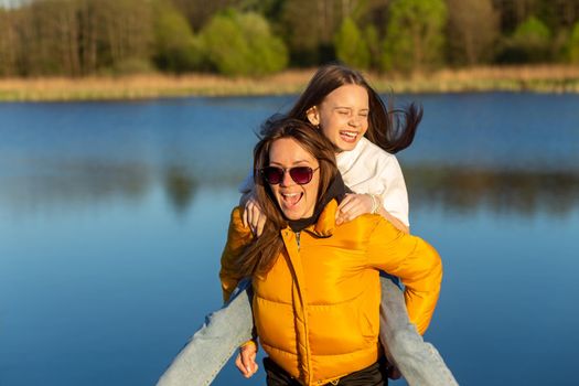 Playful mother giving daughter piggy back ride at spring lake shore. Both laughing and look happy. Spring in lake background. Closeup.