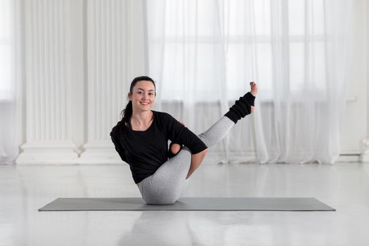 Young asian woman practicing yoga, doing variation of Navasana pose, working out, practicing yoga concept, balancing asana on gray mat in white hall.