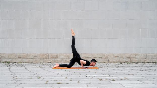 Chaturanga pose with one leg up. Fit latin man do yoga outdoor on orange mat with gray concrete wall at the background. Copy space.