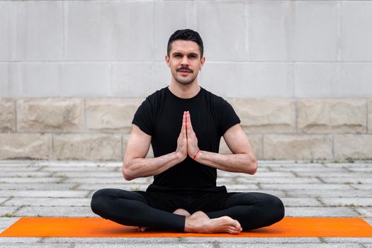 Latin man practicing yoga outdoor in a city, sitting in yoga pose on orange mat, with gray wall at the background. Front view with copy space