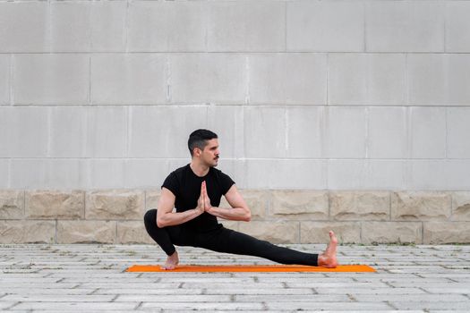 Attractive latin man doing yoga outdoors in a city. Working out exercises on orange mat with gray wall at the background.