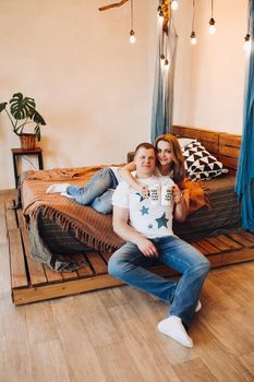 Young couple laying together on belly in bed at studio. Two people smiling and embracing, spending time together and having fun, both wearing in white t shirt. Concept of love and relationship.