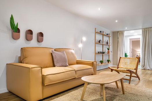 Modern boho interior of living room at cozy apartment with beige sofa, wooden table, wicker chair and stand with shelves at the background. Stylish home decor.