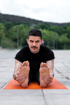 Young latin man doing yoga exercise in a park. Holding legs with hands and looking at the camera. Seated Forward Bend Yoga Pose.