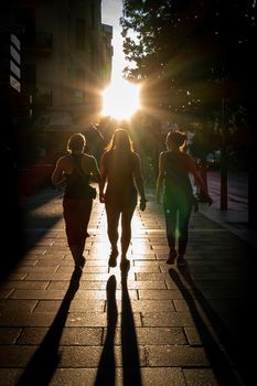 Back view of three affectionate friends walking at sunset in a city street with sun back light.