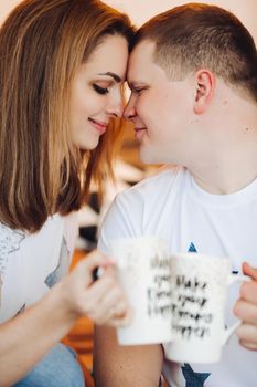 Romantic couple sitting on windows hill with lights, with creative cup in hand touching each other by heads. Boyfriend and girlfriend wearing in white t shirt and jeans embracing and enjoying in love.