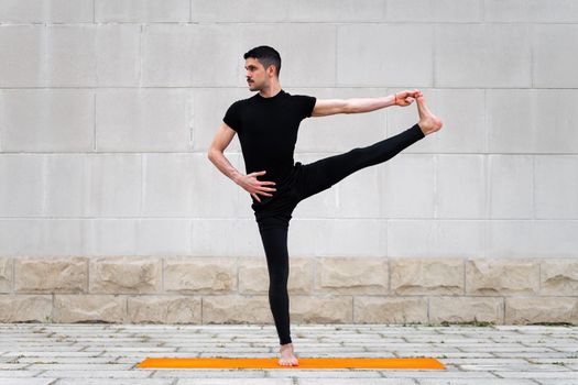 Extended Hand to Big Toe pose. Attractive latin man doing yoga outdoors in a city. Gay working out exercises on orange mat with gray wall at the background. Utthita Hasta Padangustasana, Full length