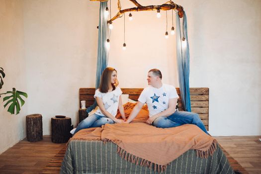 Front view of couple sitting together in bed at decorated studio. Two people smiling, looking each other, spending time together and having fun, both wearing in white t shirt. Concept of relationship.